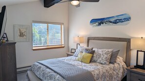 Une chambre à coucher comprenant un lit, deux tables de chevet, une fenêtre avec des stores, un ventilateur de plafond et un tableau accroché au mur.