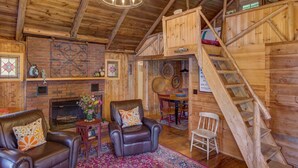 A rustic cabin interior with a fireplace, leather chairs, a wooden staircase, and a chandelier.