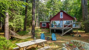 A cabin with a deck, surrounded by trees, and includes a picnic table and chairs.