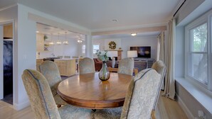 Un comedor amplio con una mesa de madera y sillas estampadas, una cocina al fondo y un ventanal con cortinas.