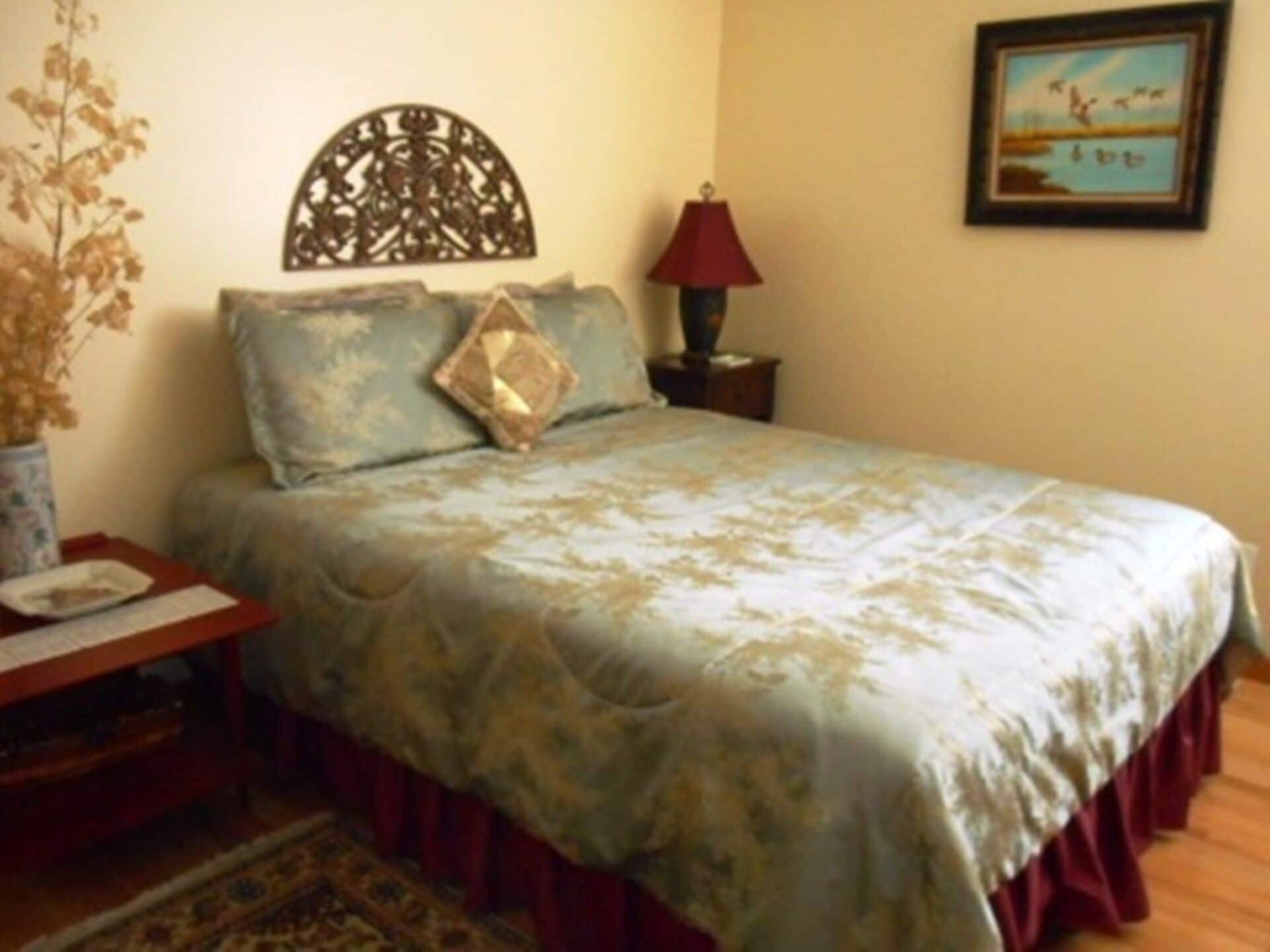 Une chambre à coucher avec un lit, une table de chevet, une lampe, un vase contenant des fleurs séchées et un tableau accroché au mur.