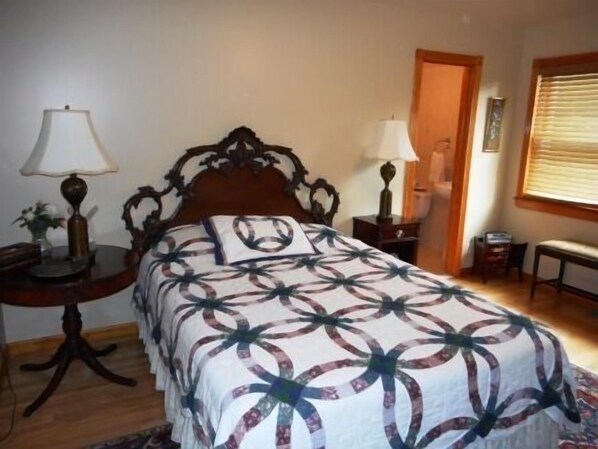 A bedroom with a wooden bed frame, a patterned bedspread, a bedside table with a lamp, a chair, and a window with blinds.
