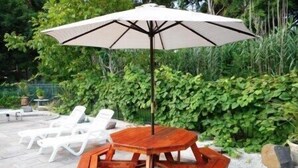 Un ensemble de mobilier de jardin en bois, comprenant une grande table et des chaises, agrémenté d’un grand parasol, le tout entouré de verdure et de chaises longues blanches.