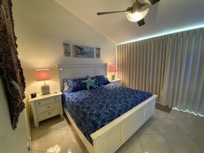 Quarto com cama, mesas de cabeceira, ventoinha no teto, obra de arte na parede e janela com cortinas.