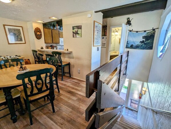 A dining area with wooden tables and chairs, a kitchen in the background, and a staircase leading to an upper level.