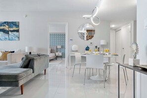 Een moderne woonkamer met een grijze bank, een eethoek met een witte tafel en stoelen, en een keuken met een wit aanrecht.