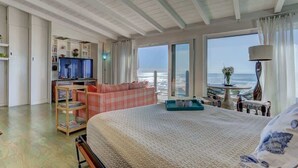 A bedroom with a large bed, a TV, a seating area with a view of the sea, and a balcony with a table and chairs.
