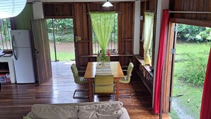 Ein gemütlicher Wohnbereich mit einer Couch, einem Holztisch und einem Esstisch mit Stühlen. Im Hintergrund ist ein Kühlschrank zu sehen, und der Raum hat Holzböden sowie einen Blick ins Grüne.