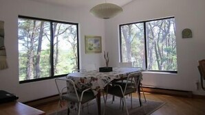 Ein Essbereich mit Tisch für vier Personen, großen Fenstern mit Blick auf einen Wald, einer Hängeleuchte und einem gerahmten Bild an der Wand.
