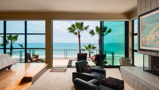 A spacious living room with a large window overlooking a beach, furnished with a sofa, chairs, and a fireplace. - LARGE CONTEMPORARY HOME ON THE SAND, HOT TUB, CHEF’S KITCHEN, ENDLESS VIEWS (Capistrano Beach)