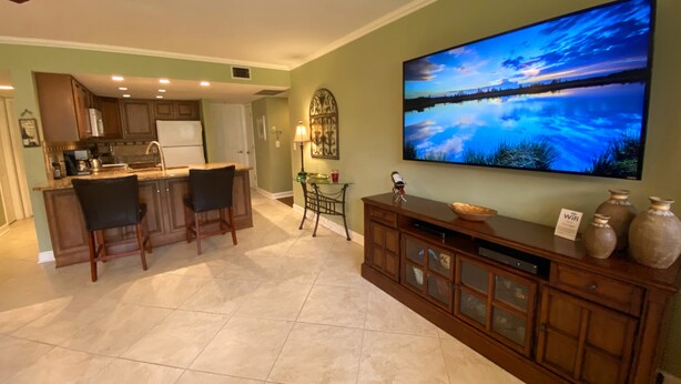 A spacious living area with a kitchen, a dining area with two chairs, a large flat-screen TV mounted on the wall, and a wooden cabinet with decorative items.