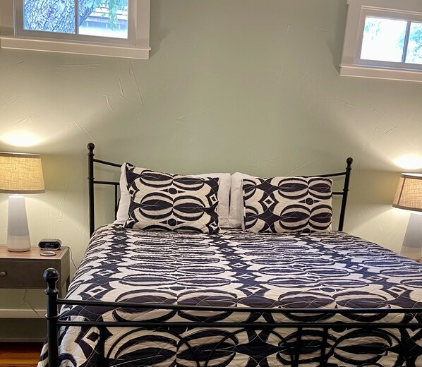 A bed with a patterned bedspread, two bedside lamps, a nightstand with a clock, and a window with a view of trees.