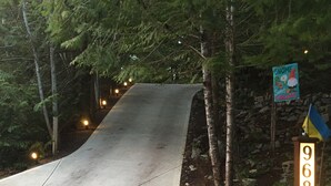Ein Betonweg führt durch ein bewaldetes Gebiet, mit einem Schild mit der Aufschrift 'GNOME Sweet GNOME' und einer Steinmauer mit einer Flagge.