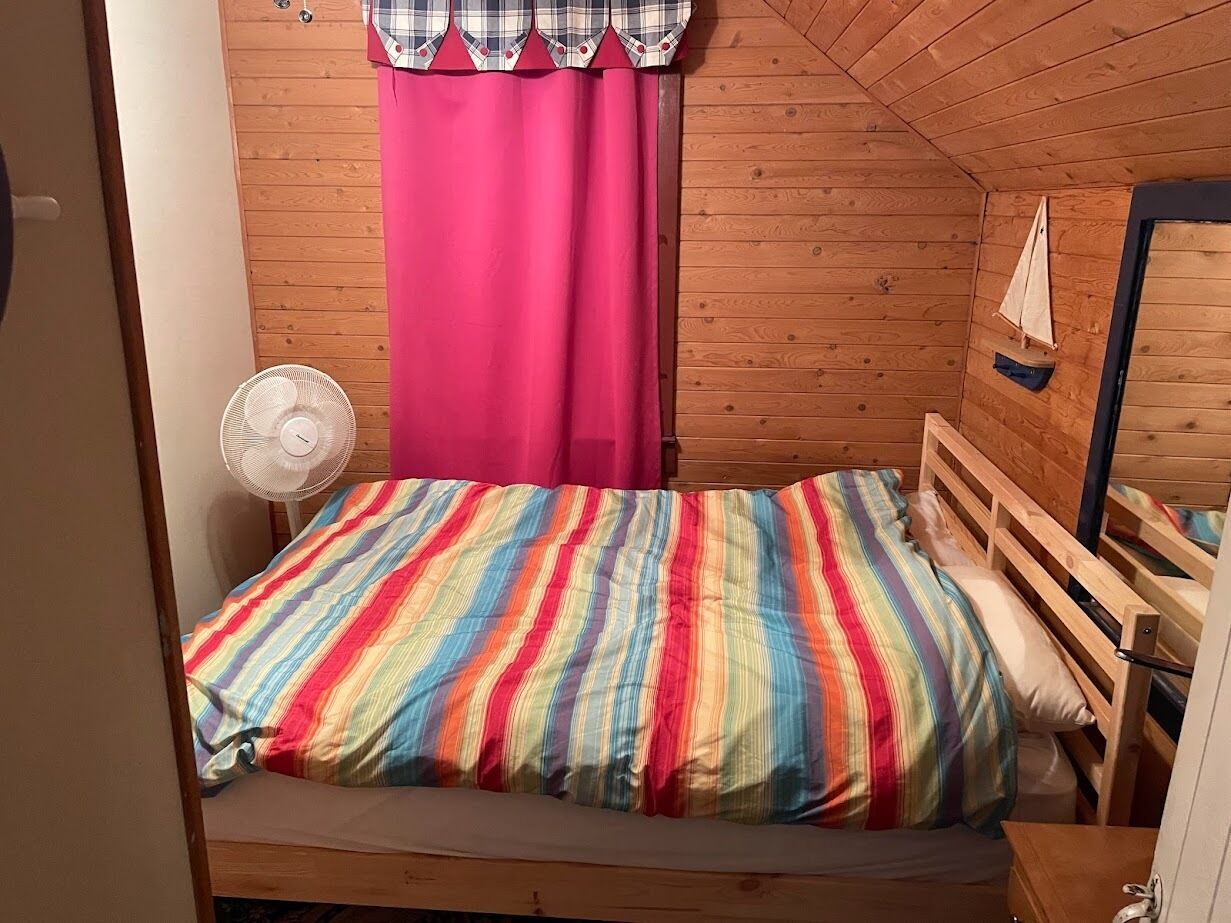 Ein kleiner, gemütlicher Raum mit Holzboden und -wänden, einem Einzelbett mit farbenfroher Streifenoptik, einem weißen Ventilator und einem rosa Vorhang.