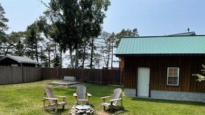 A wooden cabin with a green roof, a fenced backyard, and three Adirondack chairs around a stone fire pit.