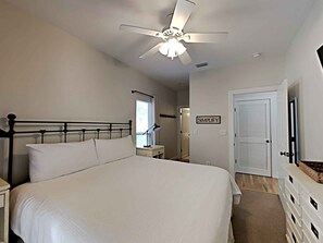 A bedroom with a large bed, a ceiling fan, a nightstand, a lamp, a door with a 'SIMPLIFY' sign, and a closet with storage drawers. - The Good Life - 100+ Top Ratings, Big Pool, Porch, 5 Min Walk to Beach, 4 Bikes (Seacrest Beach)