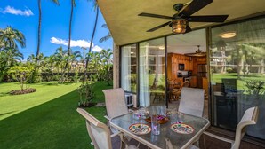 Un coin repas avec une table et des chaises en verre, un ventilateur de plafond et une vue sur un jardin luxuriant et des palmiers.