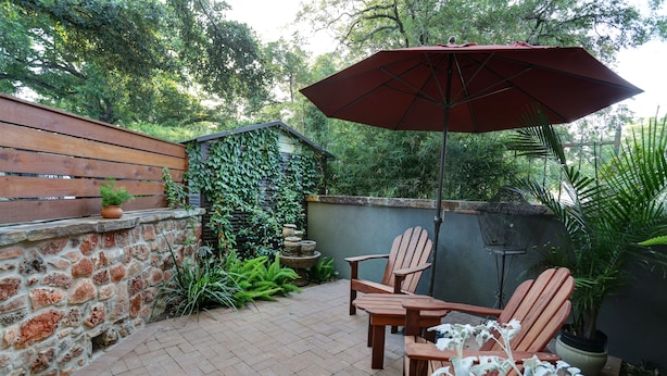 Une terrasse avec des chaises et une table en bois, un parasol rouge et un mur de pierre orné de plantes grimpantes.