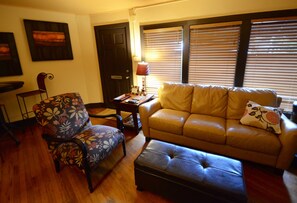 Una sala de estar con un sofá, un reposapiés, un sillón con motivos florales, una mesita con una lámpara y obras de arte en las paredes.