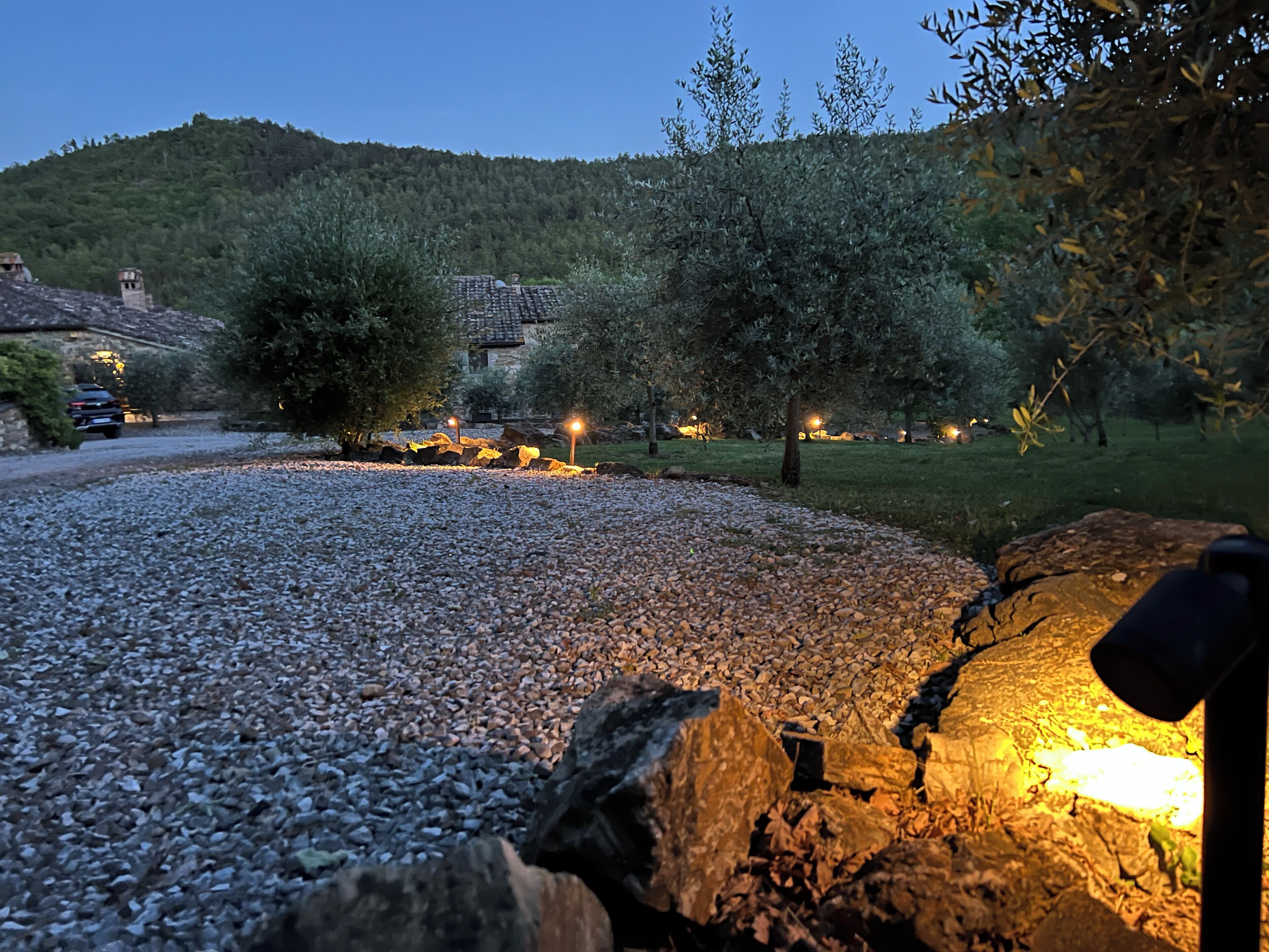 A gravel driveway leading to a stone building with a tiled roof, surrounded by trees and a hill in the background.