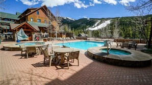Un área de piscina con sillones y mesas, un edificio de madera y montañas al fondo.