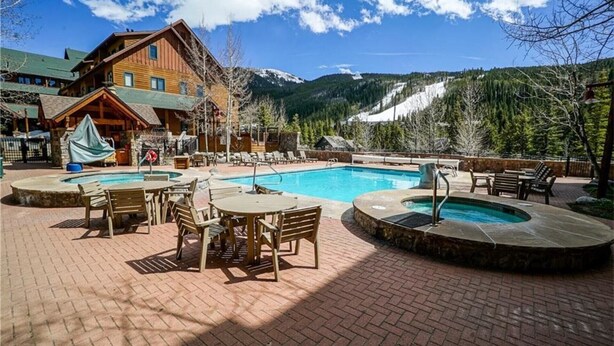 A pool area with lounge chairs and tables, a wooden building, and a mountainous backdrop. - Outstanding 2BR in River Run. Fully stocked, steps from gondola and shopping (Dillon)