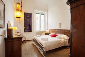 A bedroom with a bed, a desk with a lamp, a radiator, and a window with curtains.
