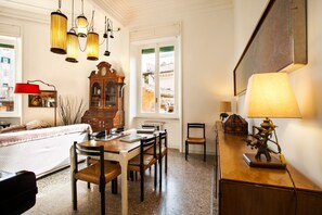 A cozy room with a wooden dining table, chairs, a red lamp, a wooden cabinet, and a large window with a view of buildings.