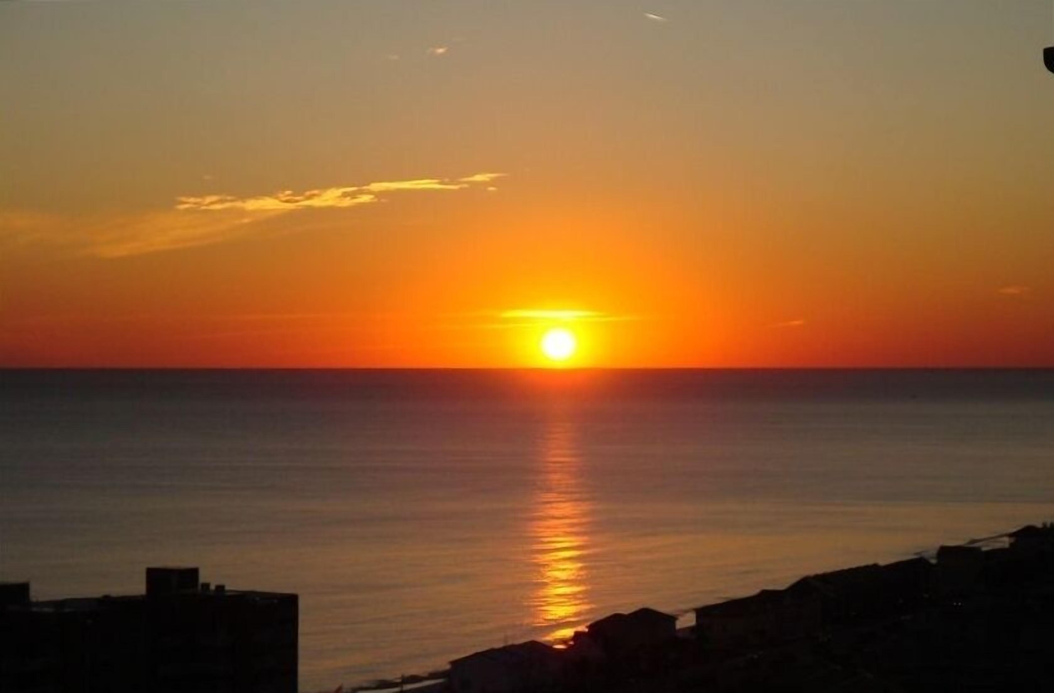 Tramonto su un mare calmo con una costa rocciosa in primo piano.