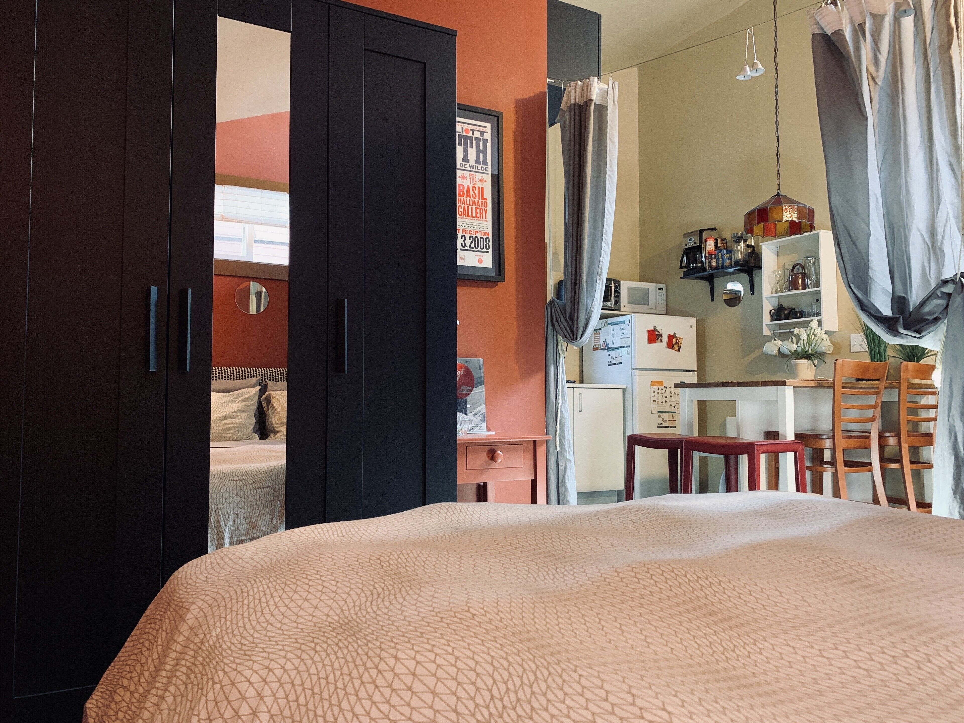 A bedroom with a bed, a wardrobe, a desk, and a kitchenette.