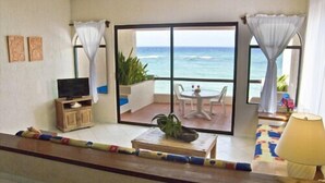 Quarto com televisão, mesa com planta e vista para o oceano por grandes janelas.