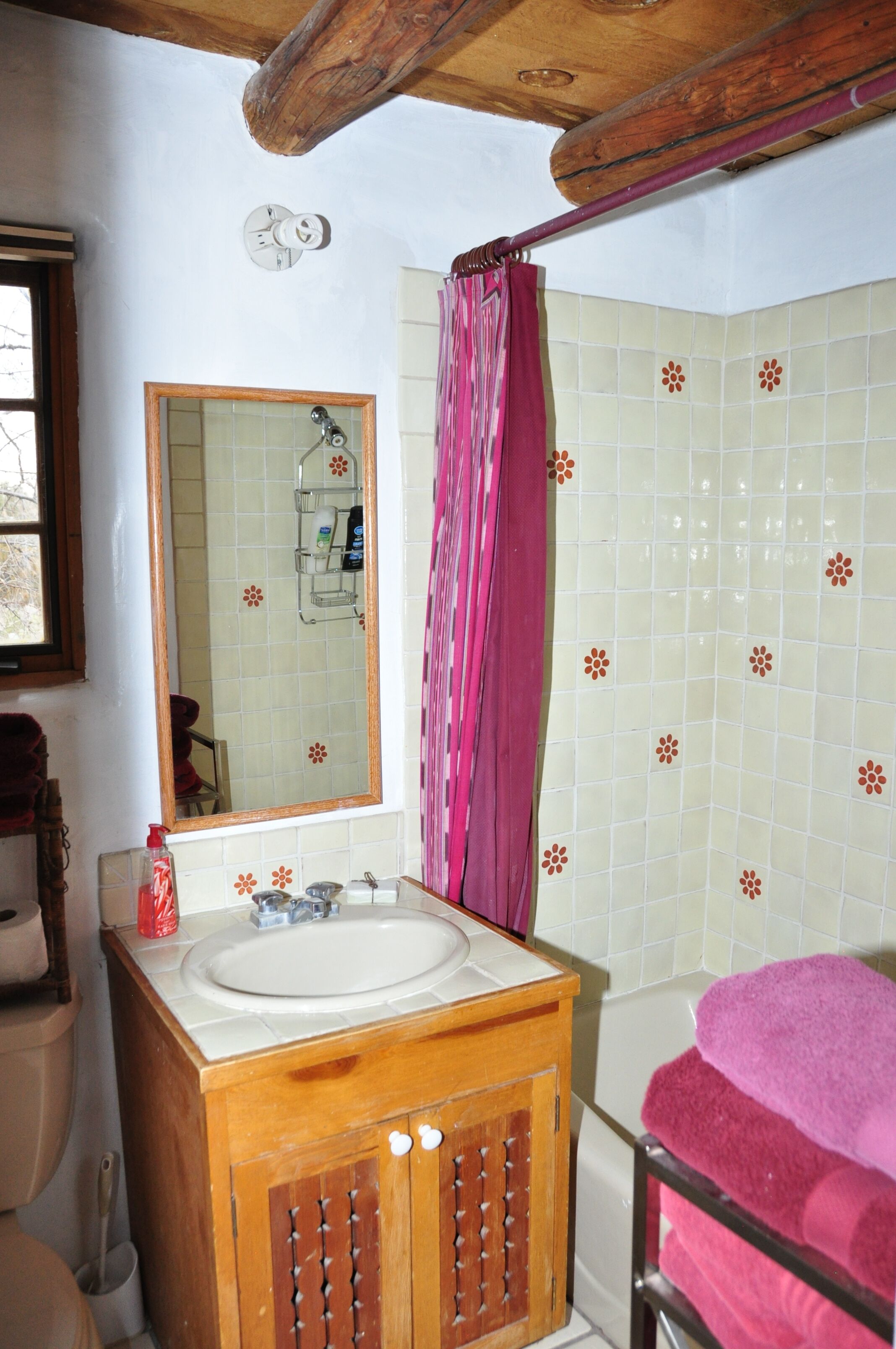A bathroom with a wooden vanity, a mirror, a sink, a shower with a pink curtain, and a tiled wall with floral patterns.