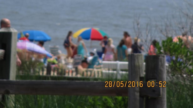 A beach scene with people under umbrellas, a wooden fence in the foreground, and a date stamp. - Only 1.5 Blocks To Ocean, 1-3 blocks of restaurants, shops, bars. Come enjoy! (Rehoboth Beach)