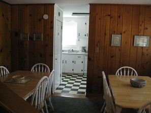 A dining room with a wooden table and chairs, a checkered floor, and a view into a kitchen with white cabinets and a window.