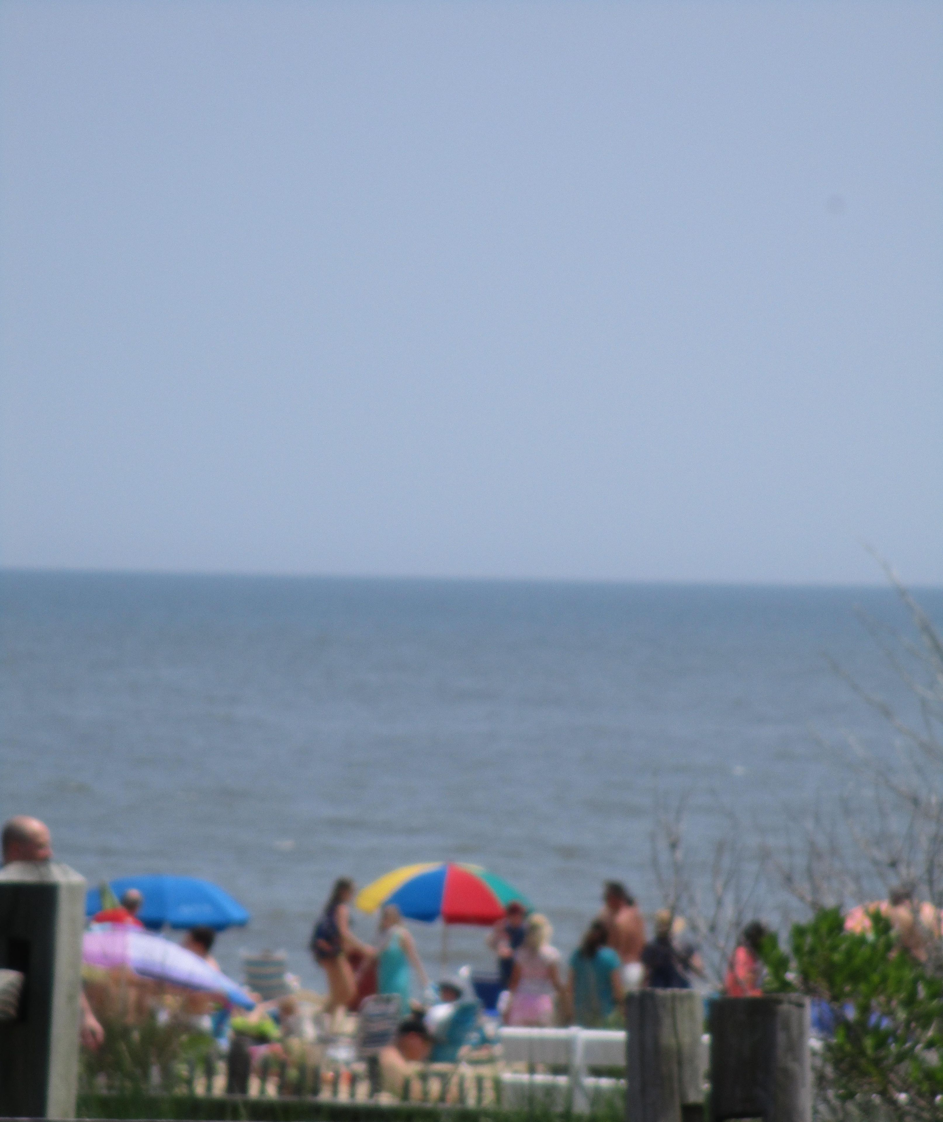 Een strandscène met mensen onder parasols, een houten hek op de voorgrond en een datumaanduiding.