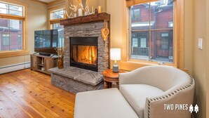 Una sala de estar acogedora con chimenea de piedra, piso de madera, un sillón cómodo y ventanas amplias que ofrecen vistas al exterior.