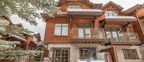 A two-story log cabin with a stone foundation, a hot tub, and a balcony with wicker furniture.