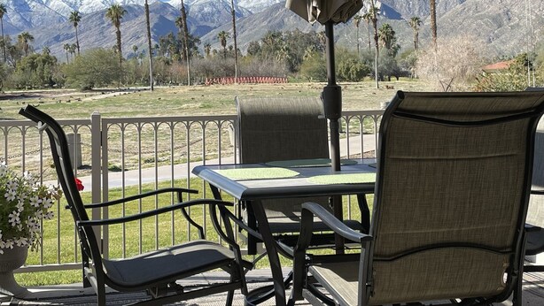 An outdoor patio with a table and chairs, overlooking a mountainous landscape with palm trees. - South Palm Springs Condo - Fantastic Mountain Views! (Palm Springs)