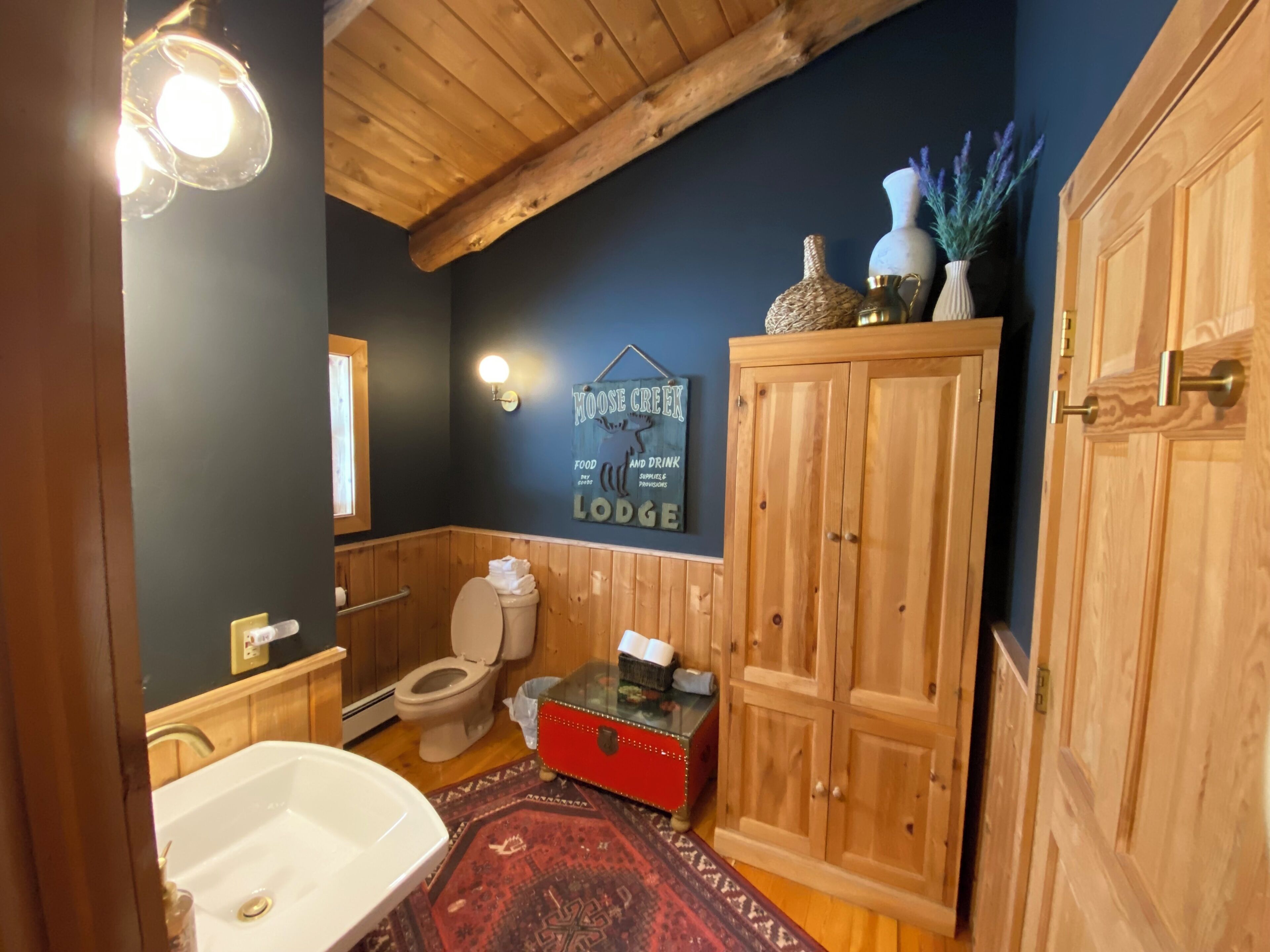 Un baño con techo de madera, un inodoro, un cofre rojo, un lavabo y una alfombra.