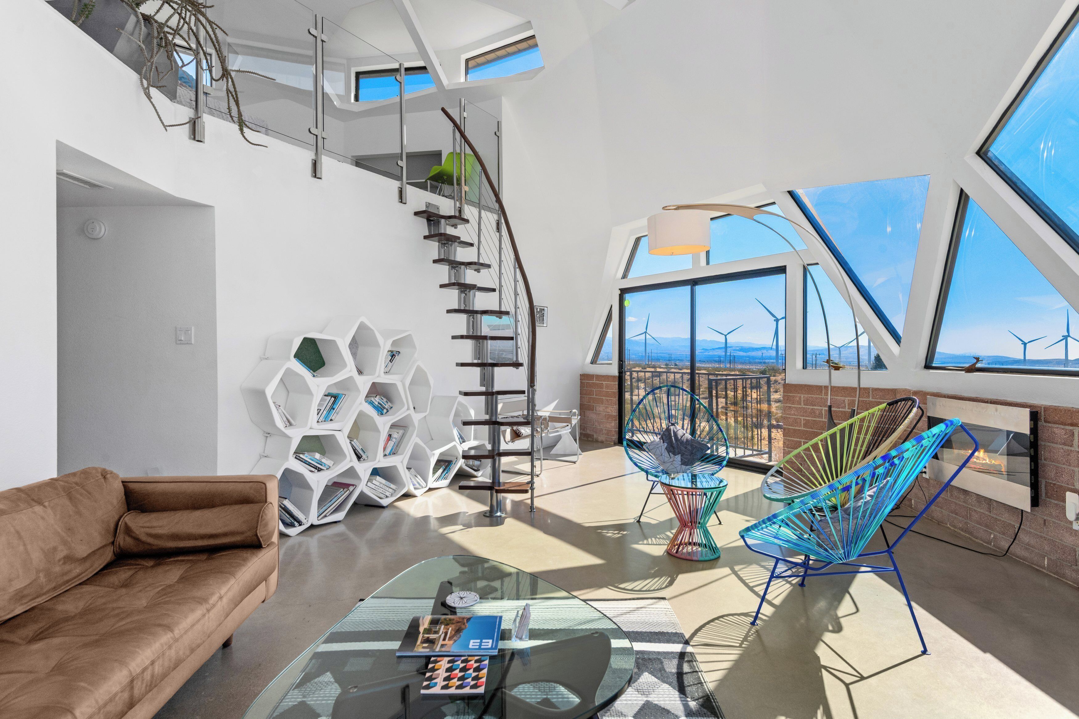A modern living room with a brown sofa, a glass coffee table, a unique hexagonal bookshelf, and a view of wind turbines through large windows.