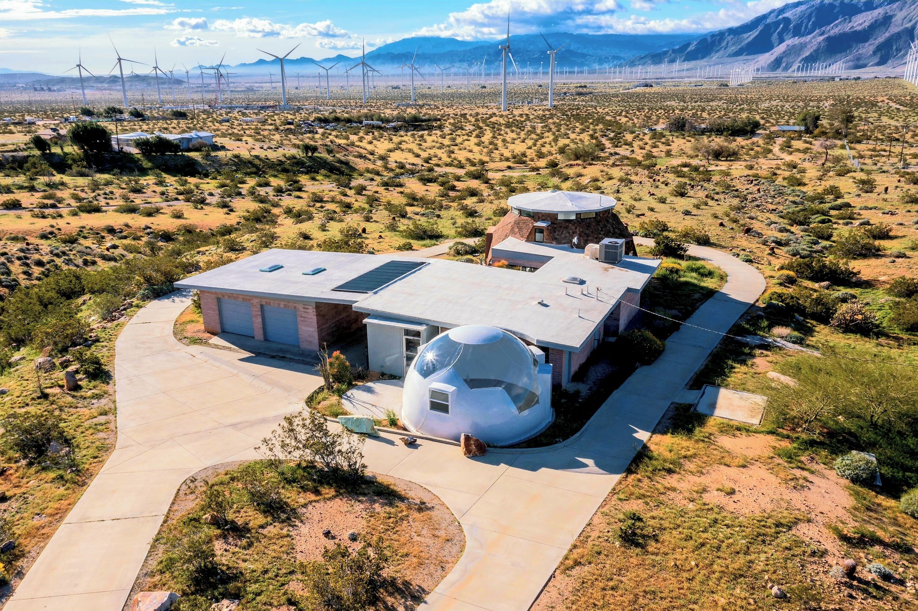 A modern building with a unique spherical structure, surrounded by a desert landscape and wind turbines in the background.