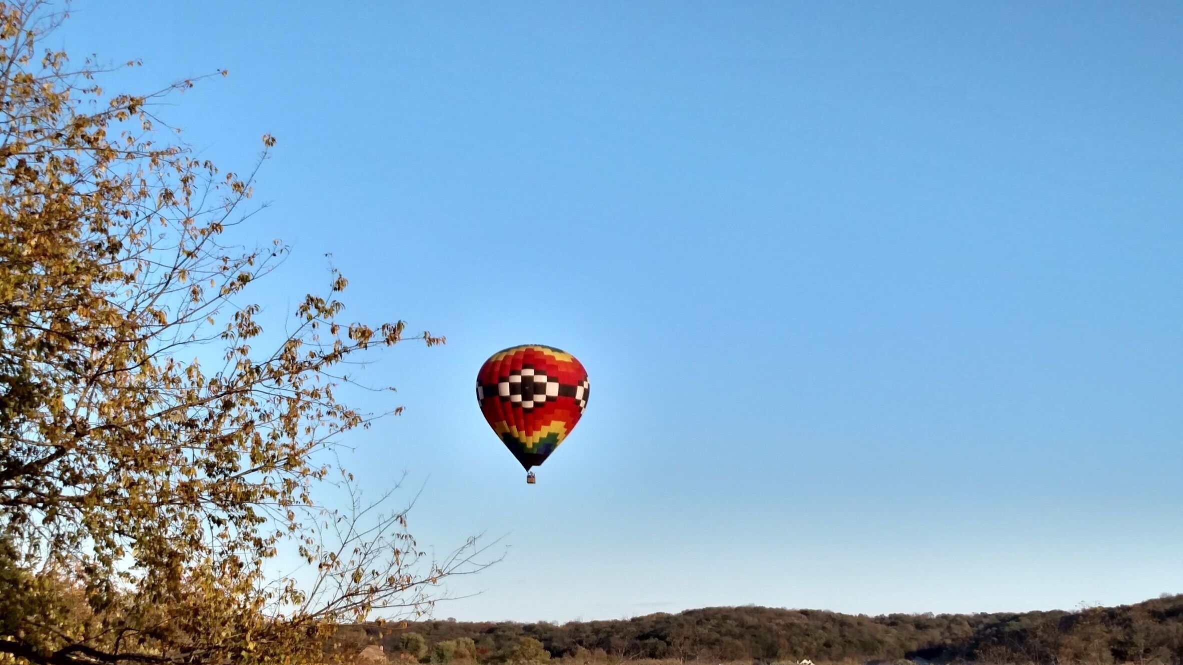 A hot air balloon with a checkered pattern flying over a landscape with trees and hills.