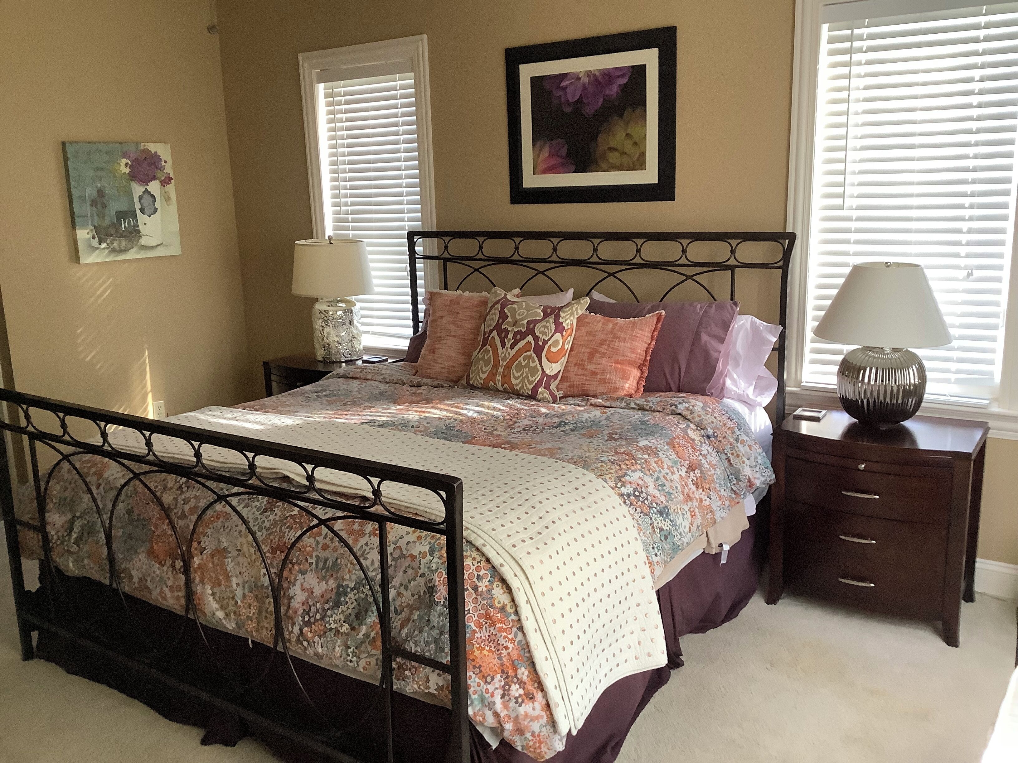 A bedroom with a bed, two bedside tables, a nightstand, a lamp, a framed picture, and a window with blinds.