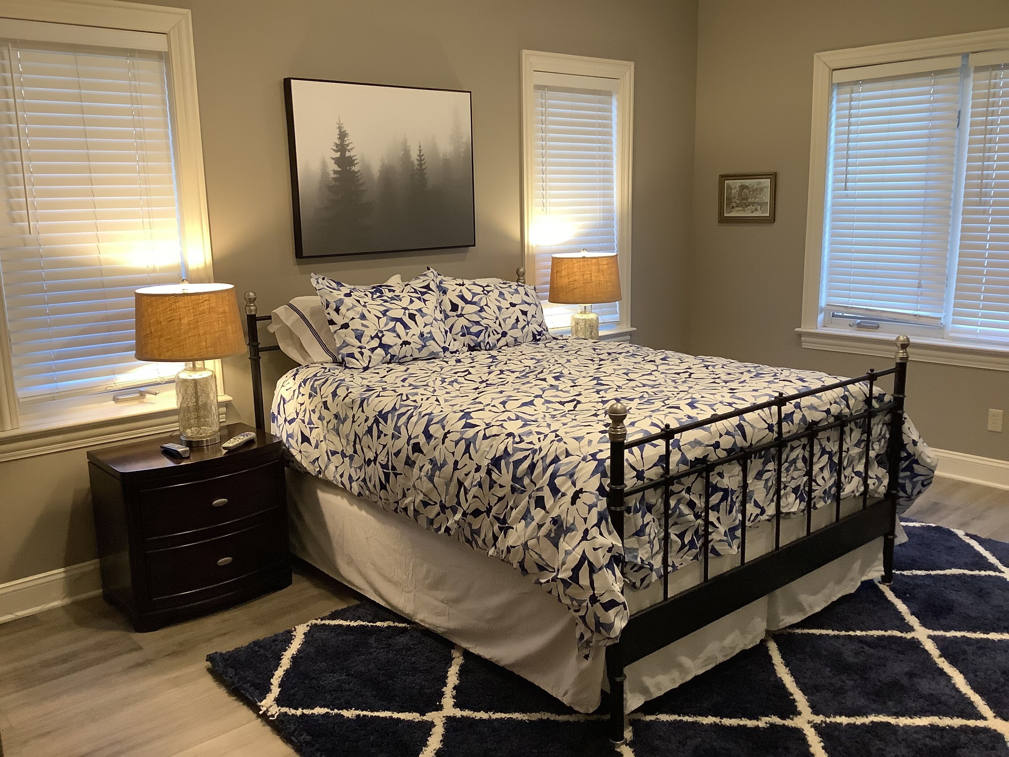 A bedroom with a bed, two bedside tables, a nightstand, a rug, and two windows with blinds.