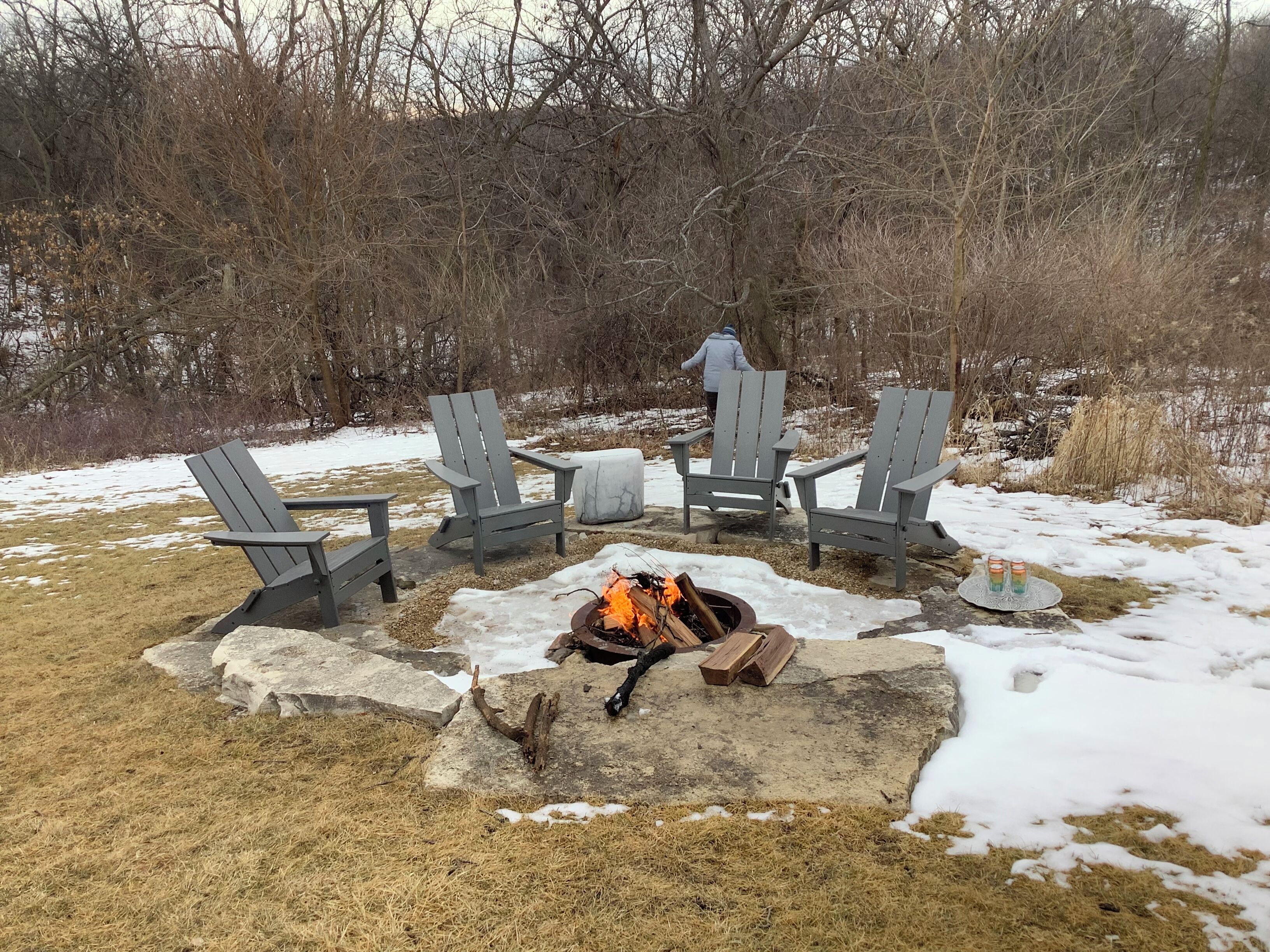 A fire pit with logs and a table with chairs around it, set in a snowy outdoor area with trees in the background.