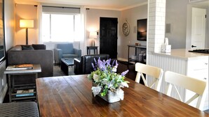 Ein Wohnzimmer mit einer Couch, einem Esstisch mit Blumenvase und einer Küchenzeile inklusive weißem Schrank und Wanduhr.