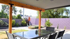 Eine überdachte Terrasse mit einem Glastisch und Stühlen, umgeben von einer violetten Ziegelwand und Pflanzen.