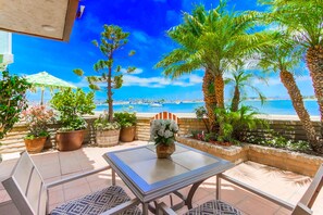 An outdoor patio with a table and chairs, surrounded by potted plants and palm trees, overlooking a beach with boats.