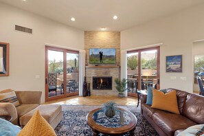 A spacious living room with a fireplace, a large flat-screen TV, and a view through glass doors to an outdoor patio.
