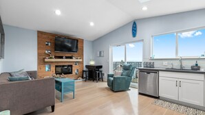A modern living room with a fireplace, a sofa, a teal armchair, and a kitchen area with a dishwasher and cabinets. - Coastal View Home - Newly Remodeled.  Water Taxi Ride Away to see the WORLD CUP! (Seattle)